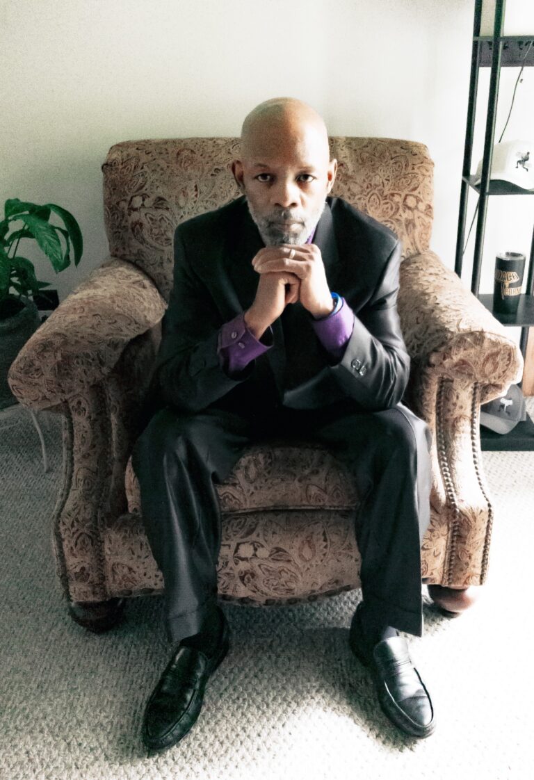 Coach T seated in an armchair in a dark suit, hands folded